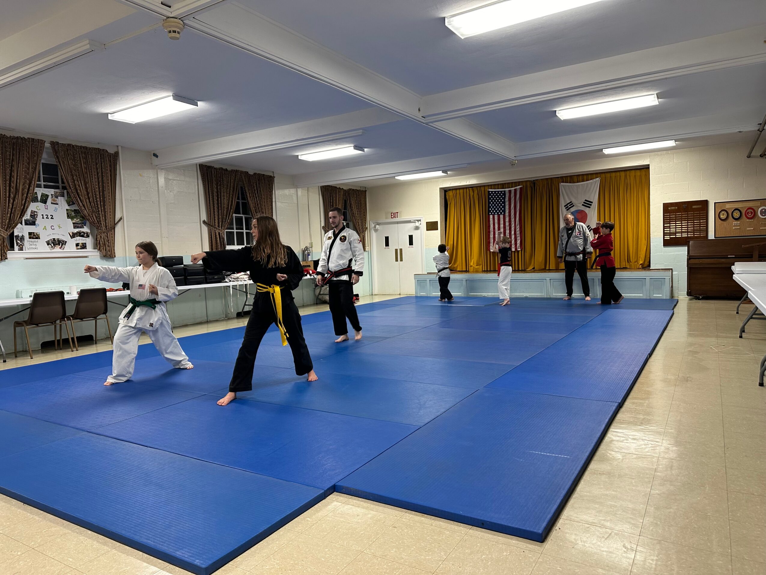 People practicing martial arts on blue mats in a room with yellow curtains and an American flag, some wearing uniforms.
