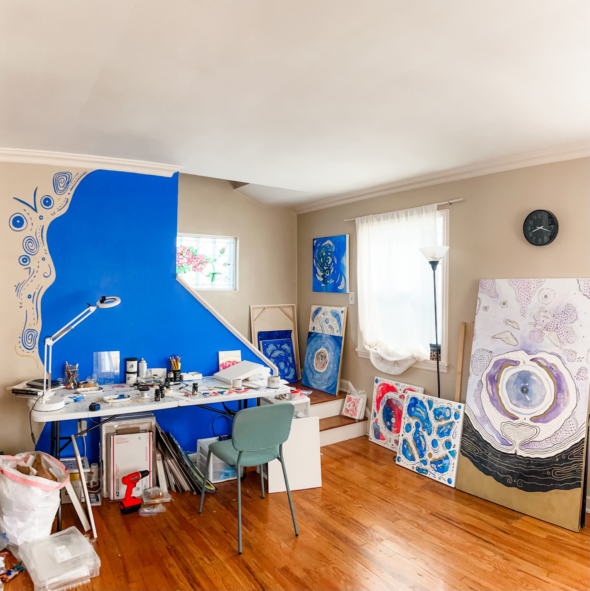 Room with blue and white wall, desk, chair, paintings, window, and wooden floor.