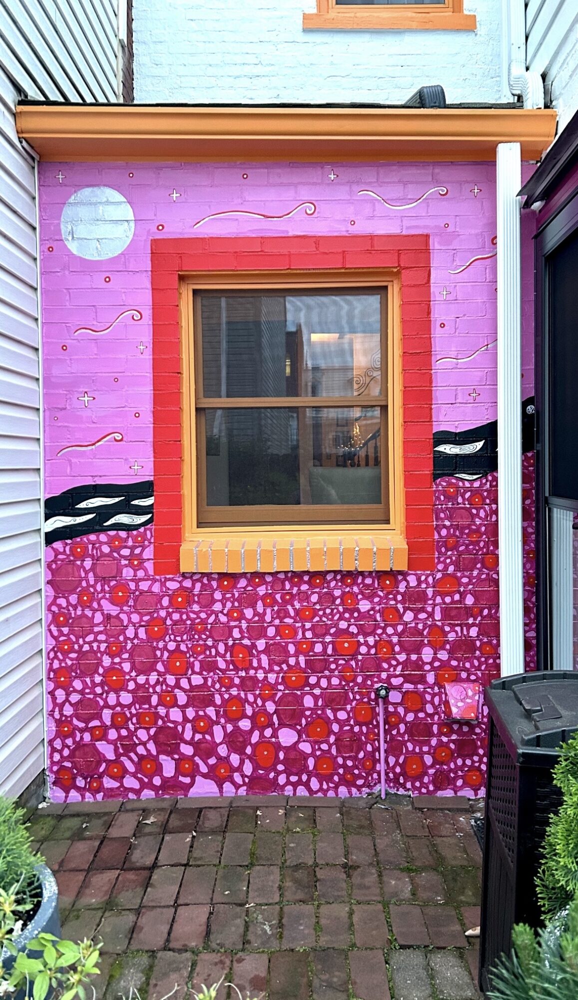 Colorful mural on a brick wall with a window, featuring pink, purple, and orange patterns, and a small outdoor area.