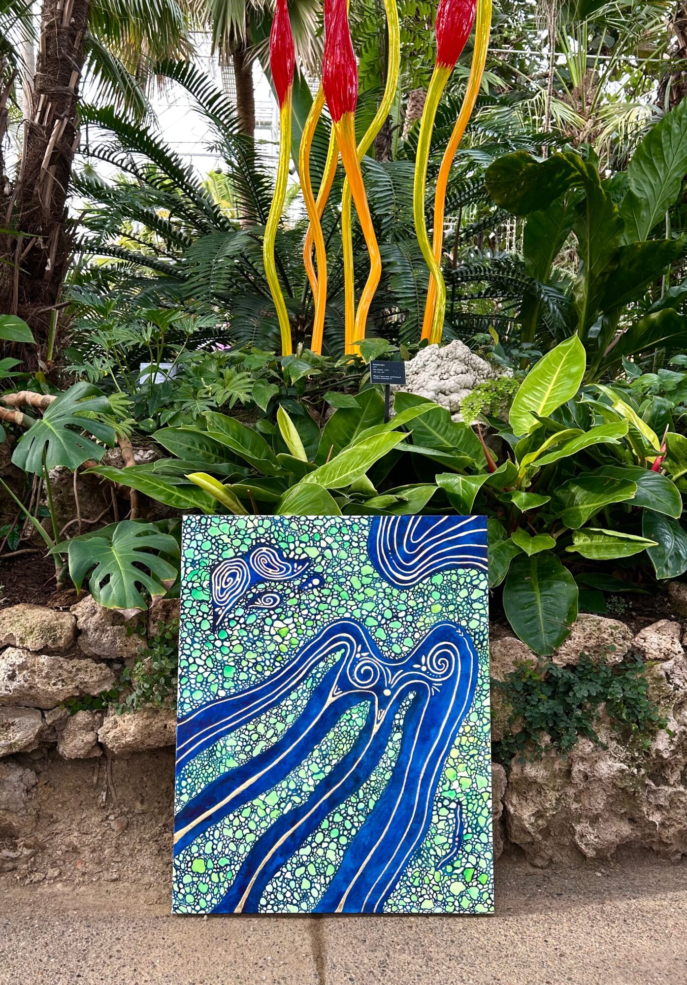 Colorful abstract painting with blue, green, and white patterns, placed outdoors among lush green plants and rocks.