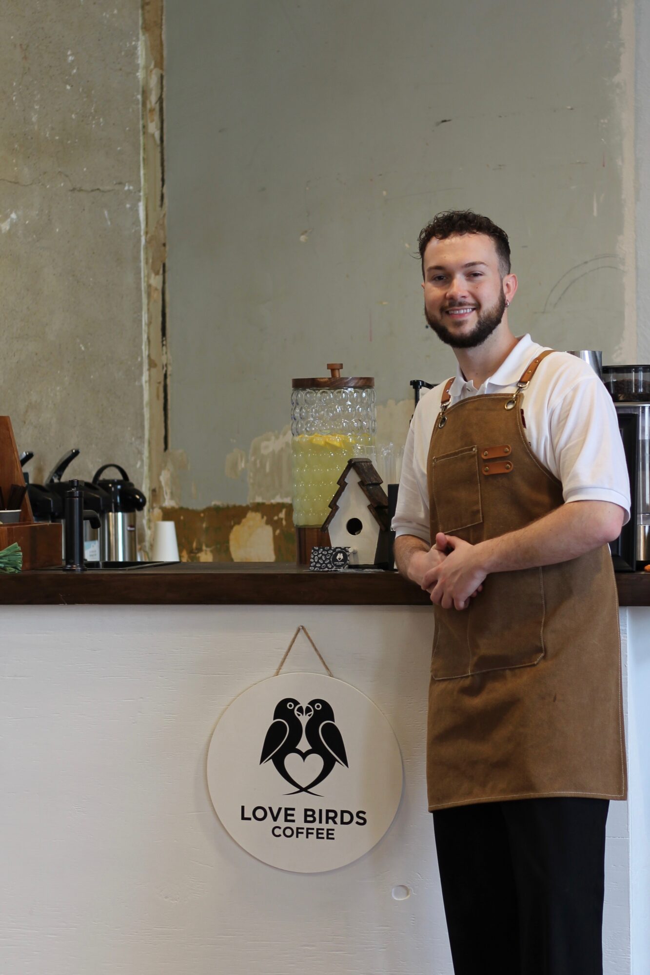 Man with a beard and white shirt wearing a brown apron stands behind a coffee counter, smiling. Sign reads 'Love Birds Coffee'.
