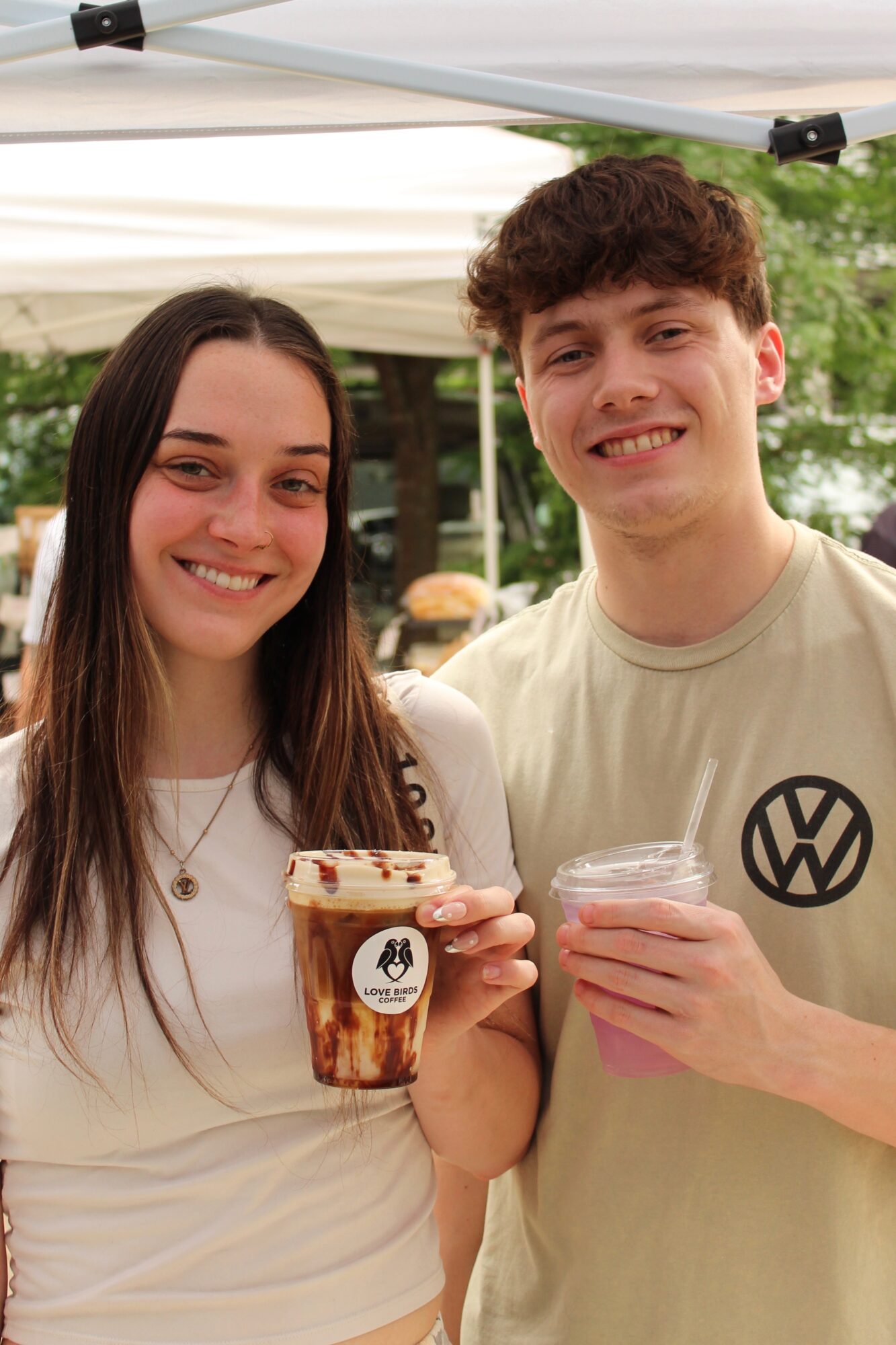 Two smiling young people holding drinks outdoors under a canopy, with trees in the background.