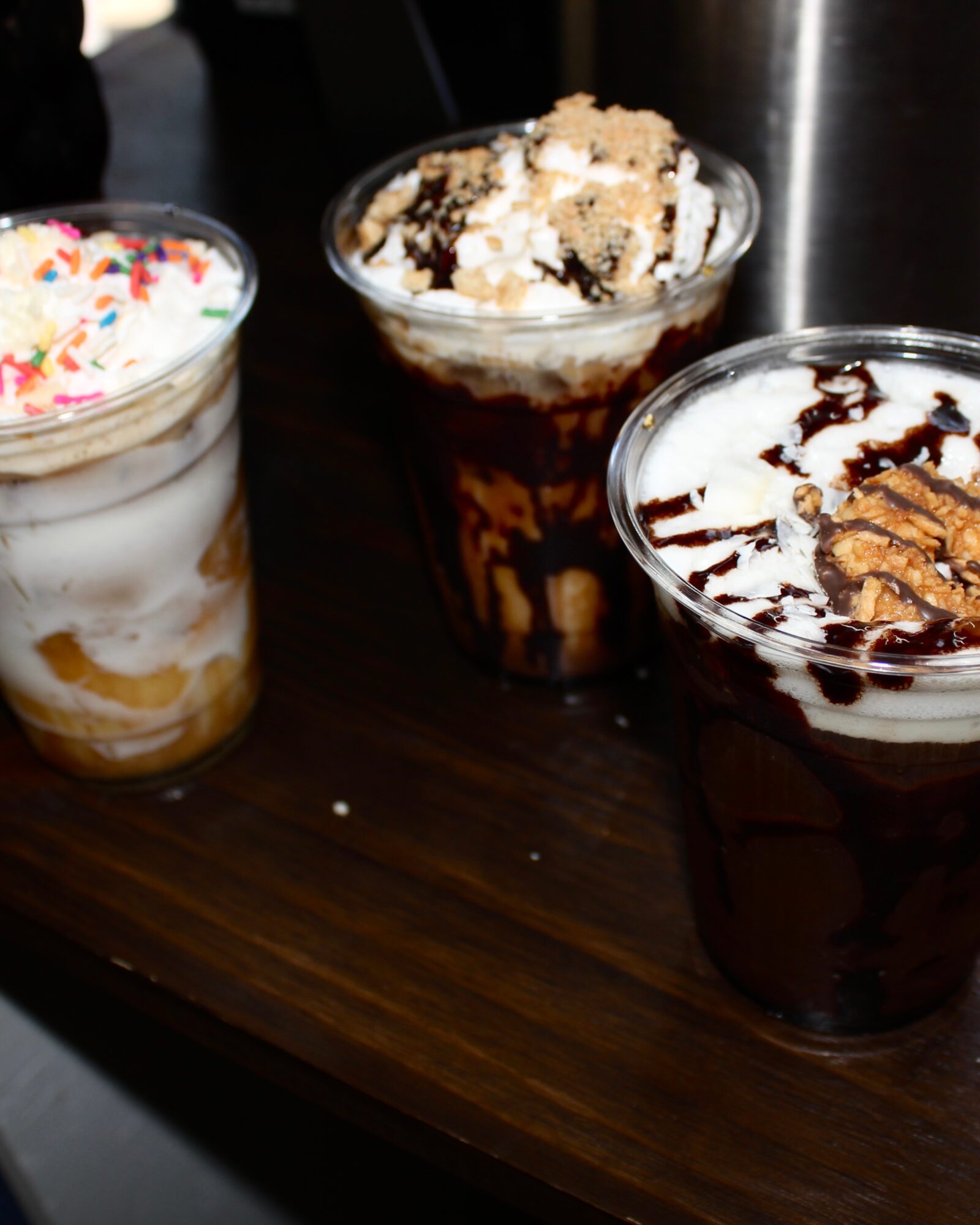 Three dessert drinks with whipped toppings and toppings on a dark wooden table.