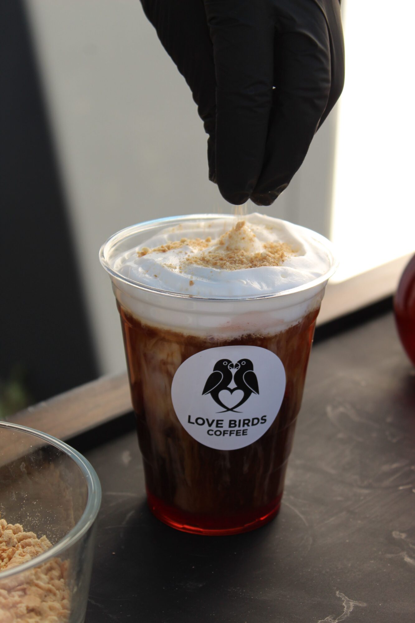 Person wearing black glove holding a plastic cup with a drink and whipped cream, on a black surface.