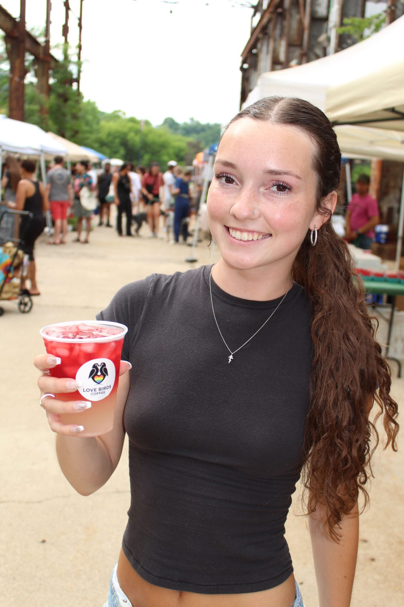 Young woman with long curly hair smiling, holding a red cup outdoors, wearing a black shirt, in a busy outdoor setting.