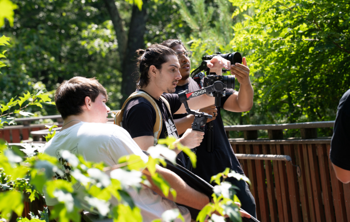 Group of people outdoors, one person holding a camera, surrounded by green trees and foliage.