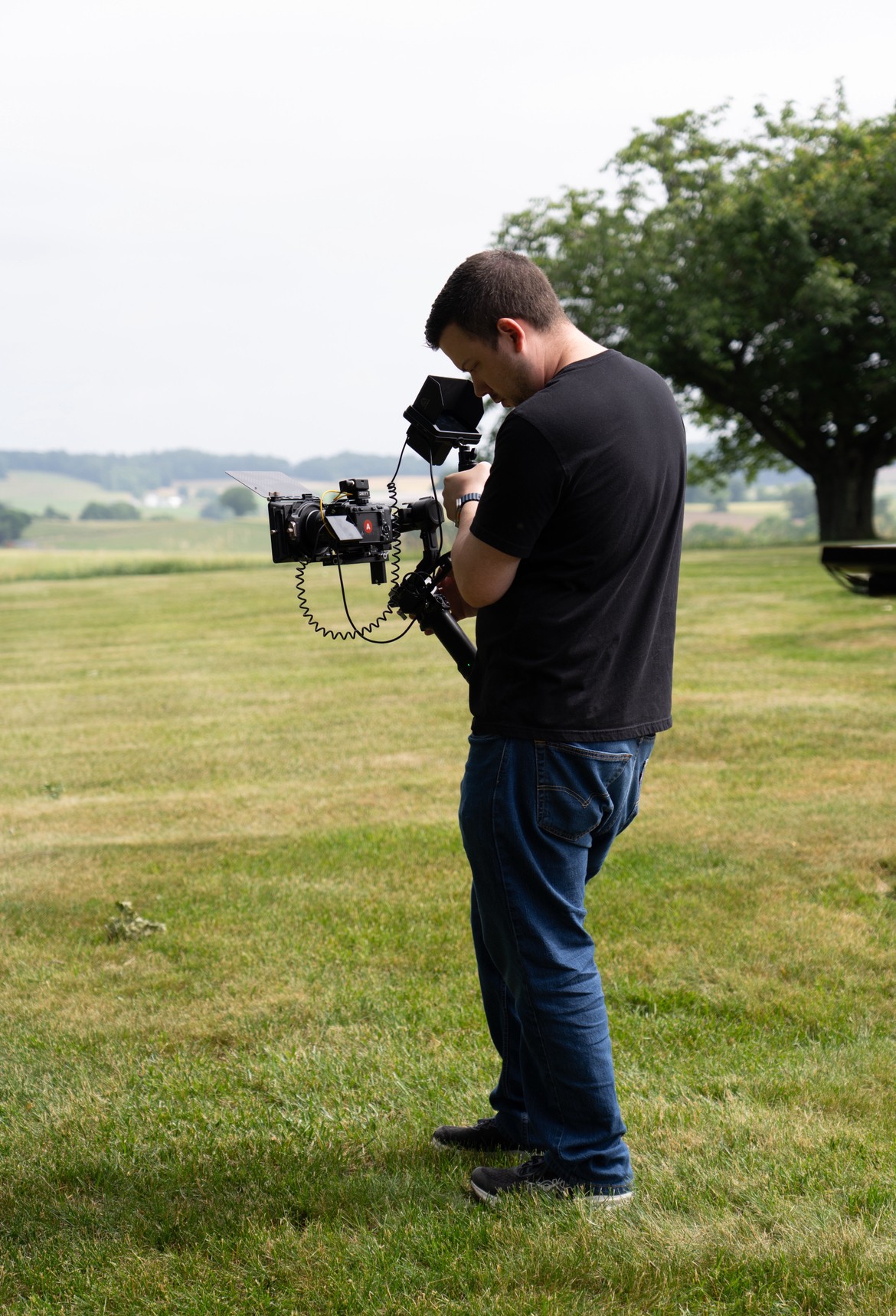 Man operating a professional camera outdoors on a grassy field with trees in the background.