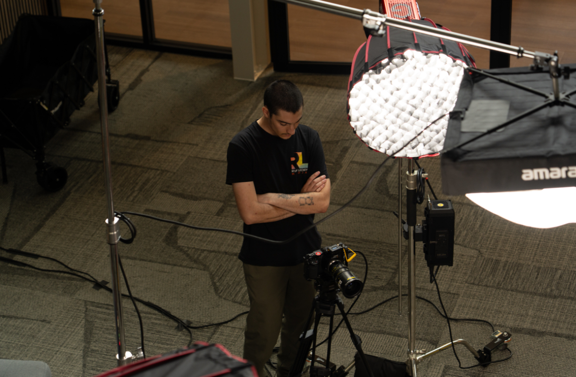 Person standing in a studio with lighting equipment and camera, arms crossed, wearing a black shirt and glasses.