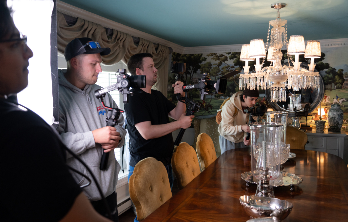 Group of people filming in a room with a chandelier and a long wooden table, some chairs, and a mural on the wall.
