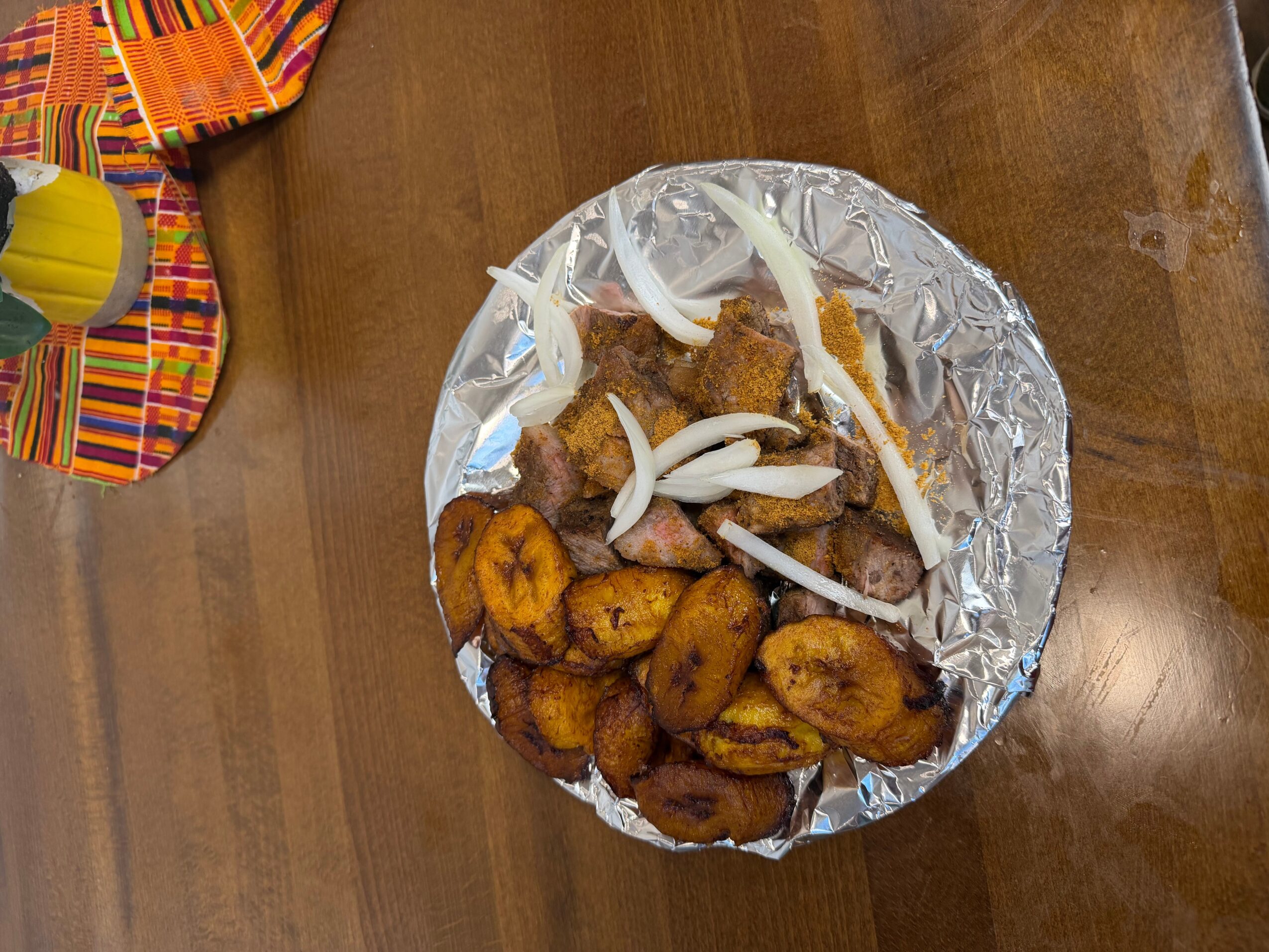 Fried plantains and grilled sausages on a foil-covered plate, garnished with sliced onions, on a wooden table.