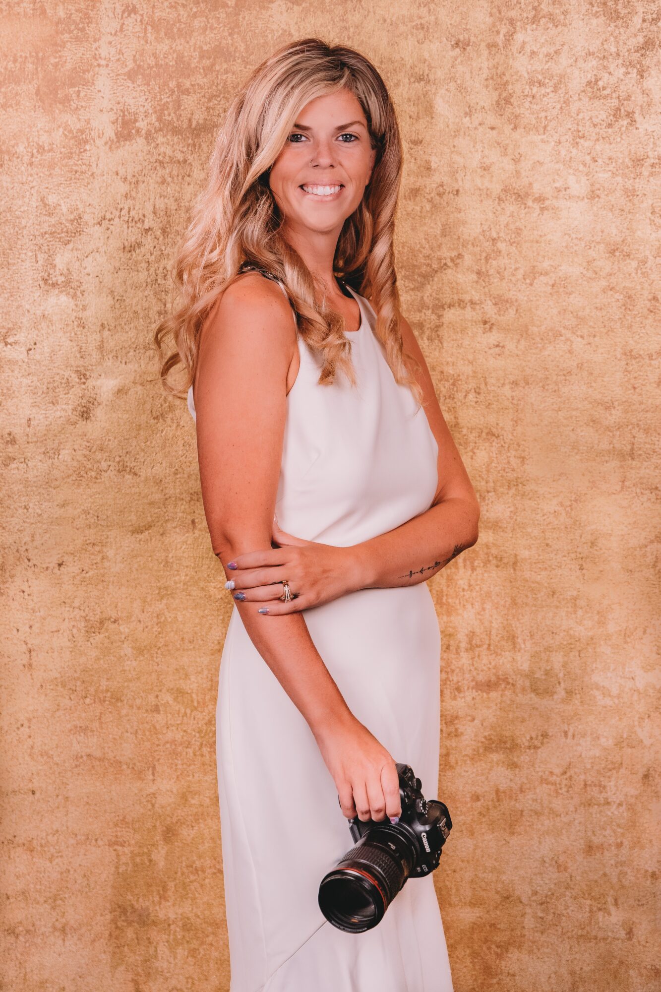 Woman with long wavy hair in a white dress holding a camera, smiling, standing against a textured beige background.