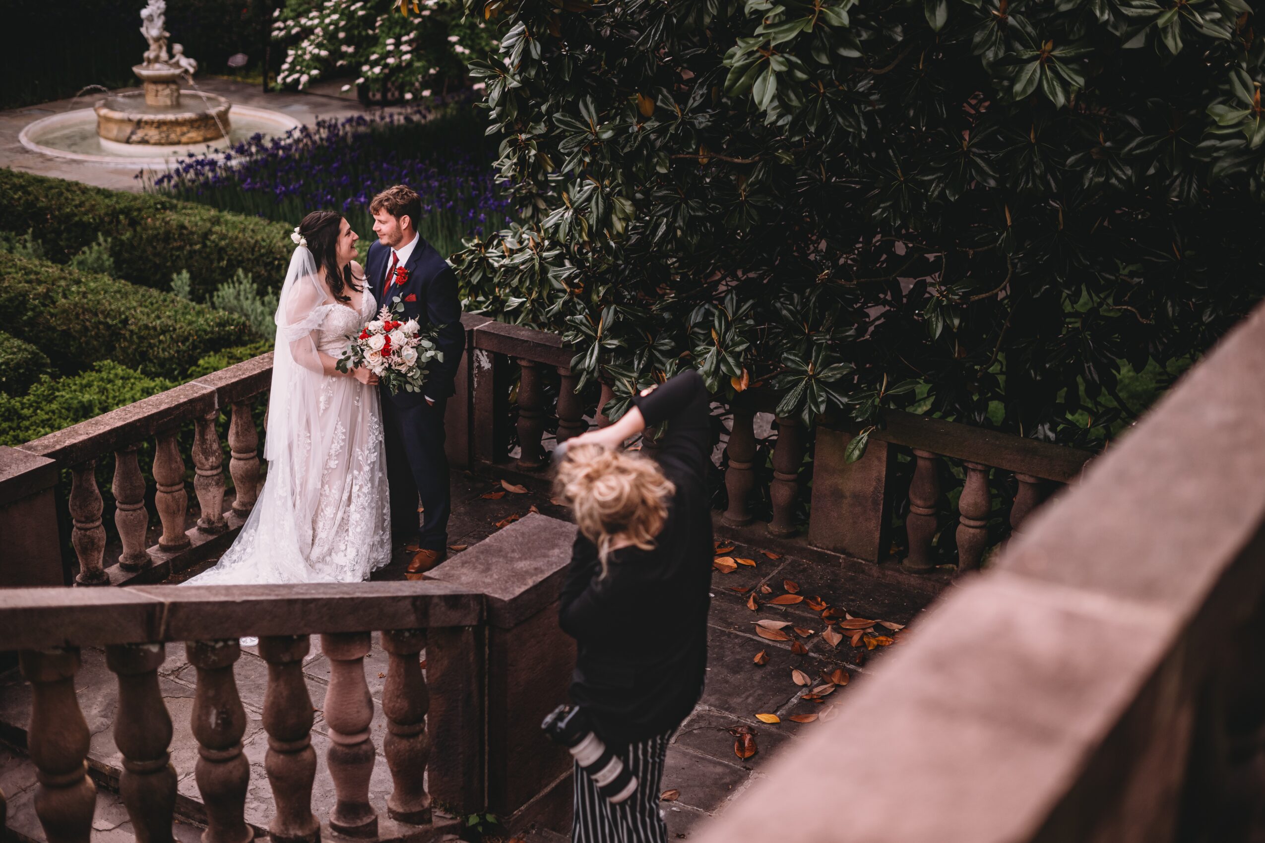 A bride and groom stand on a stone balcony, holding hands and looking at each other, with a person taking a photo nearby.