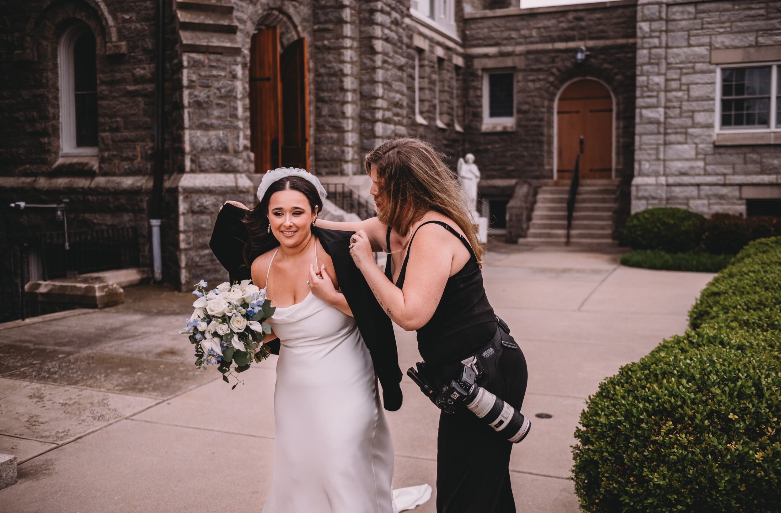 A bride in a white dress holding a bouquet, smiling, with a photographer adjusting her dress outside a stone building.