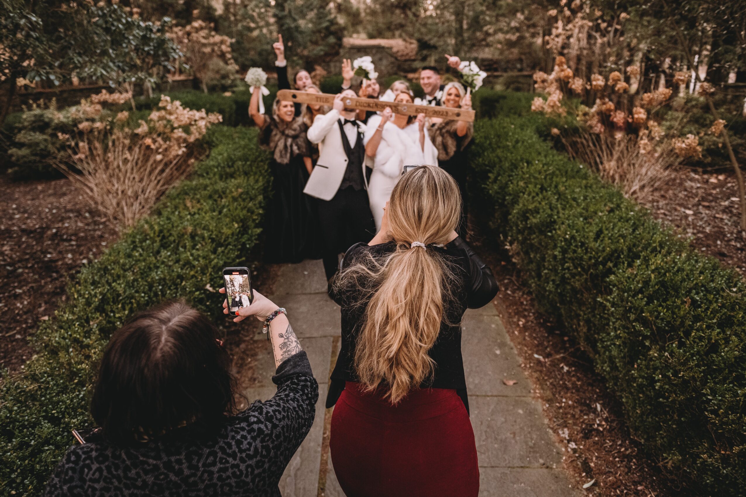 Group of people taking a photo in a garden with bushes and flowers, some holding drinks, during daytime.