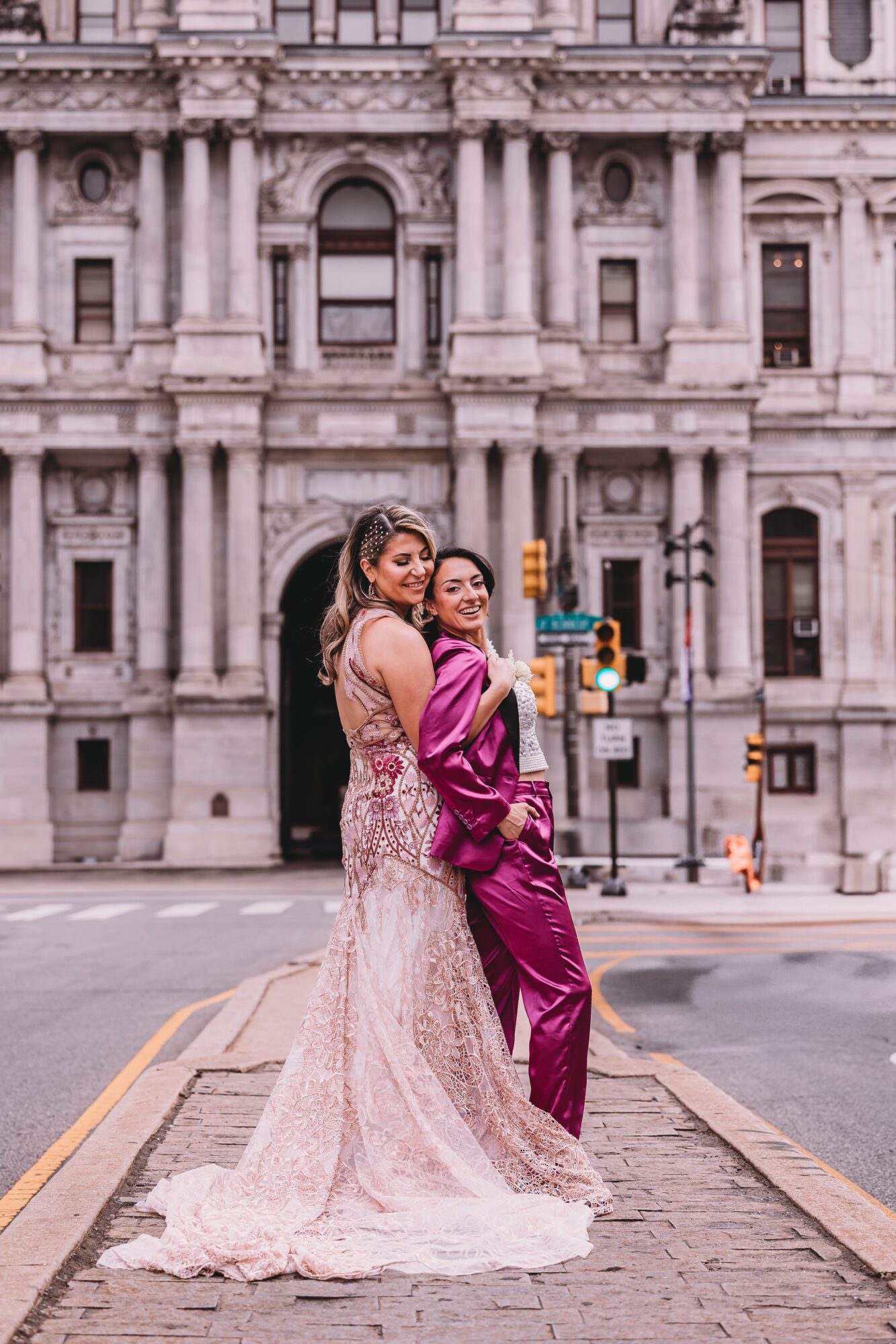 Two women in elegant dresses stand on a city street in front of a historic building, smiling and embracing.