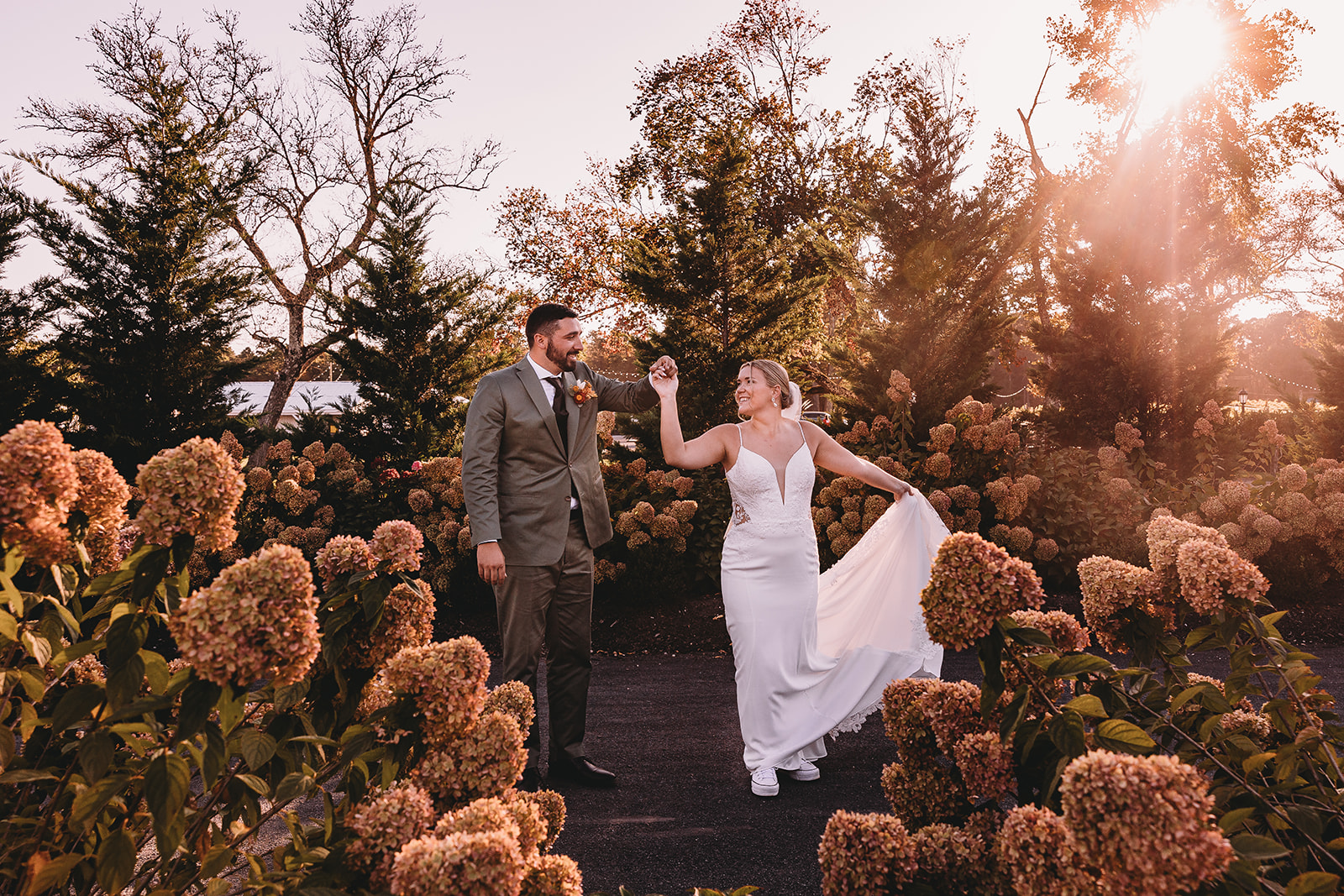 A bride and groom stand outdoors among flowers and trees, with sunlight shining behind them, celebrating their wedding.