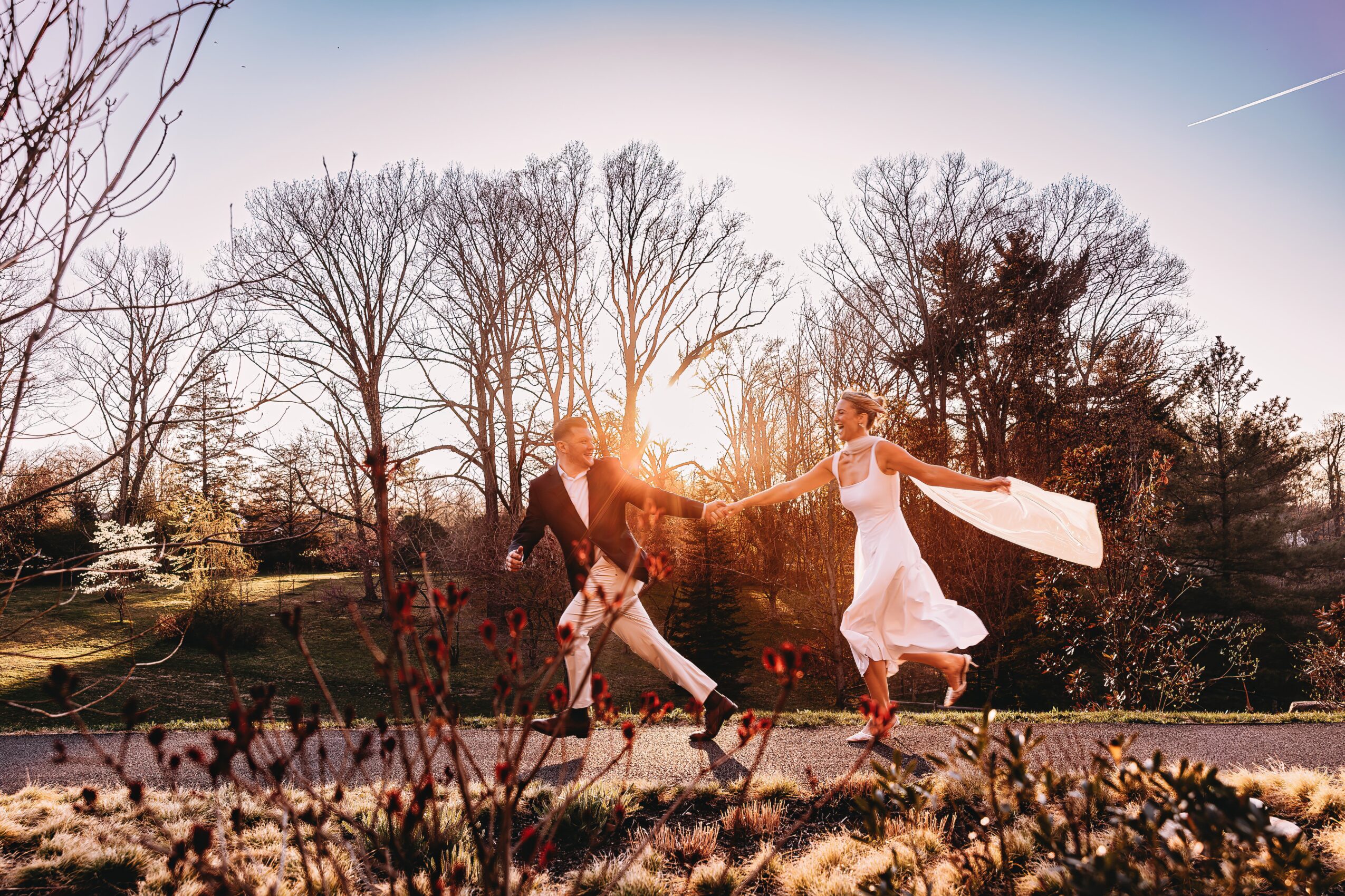 A couple holding hands and dancing outdoors during sunset, with trees and a clear sky in the background.