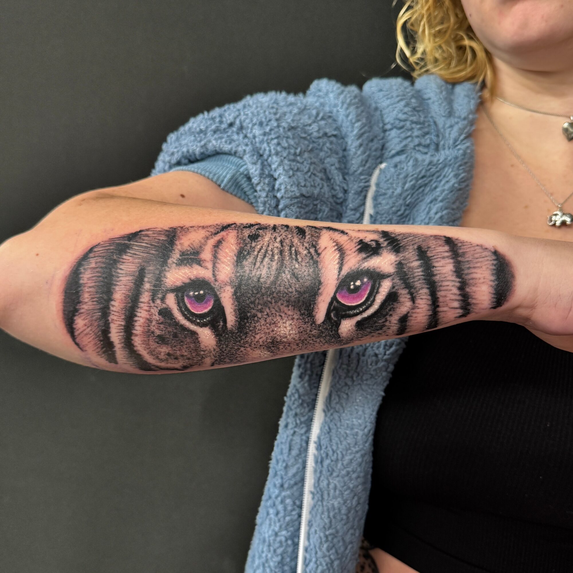 Person with a tiger face tattoo on their forearm, showing pink eyes and striped fur pattern, wearing a blue hoodie and black top.