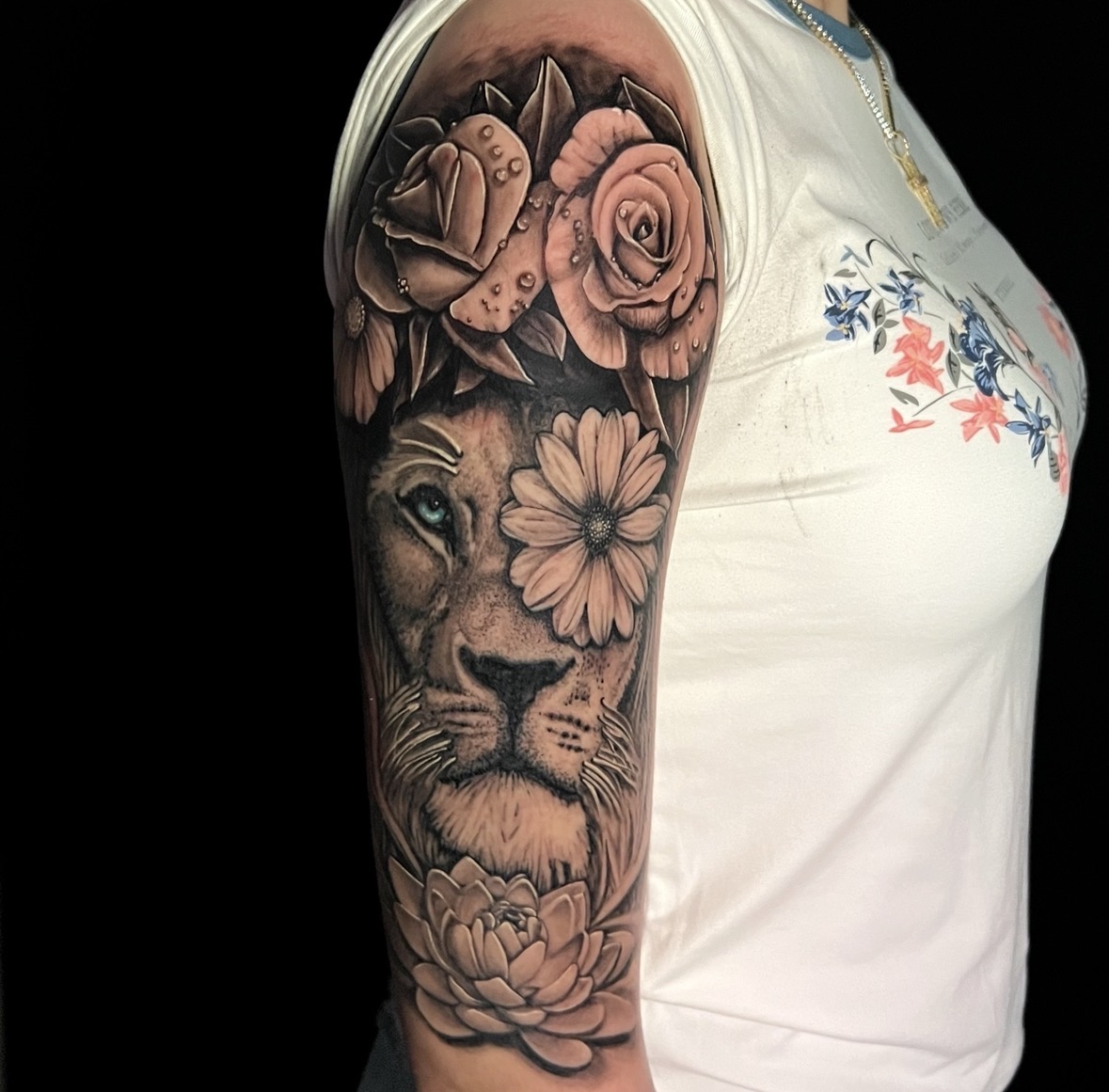 Tattoo of a lion's face surrounded by roses and flowers on person's upper arm, with part of a white shirt visible.
