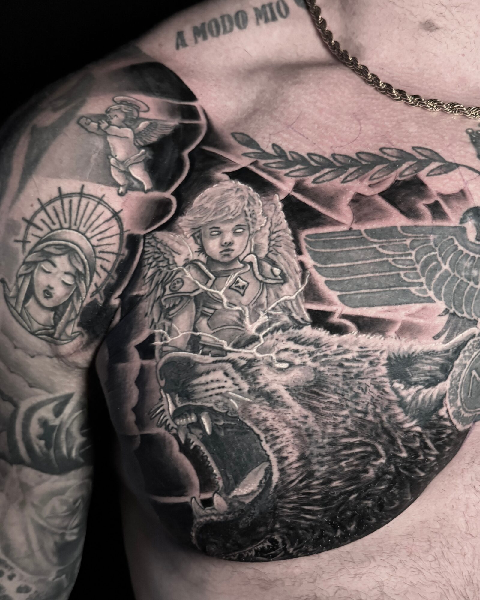 Tattooed chest with a girl in armor, a winged creature, clouds, and a sun with a face, in black and gray ink.