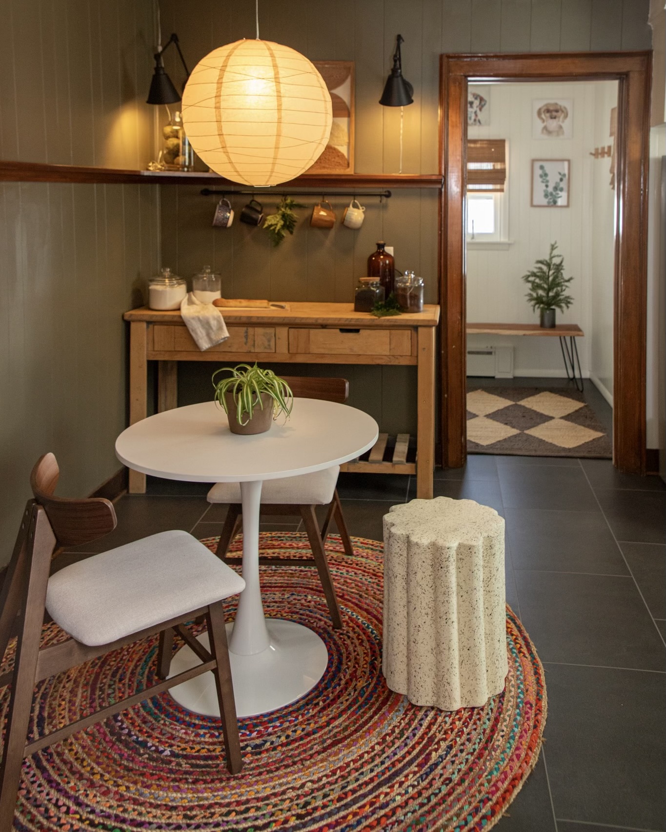 Interior of a cozy room with a round white table, a wooden chair, a colorful rug, and a doorway to another room with a plant.