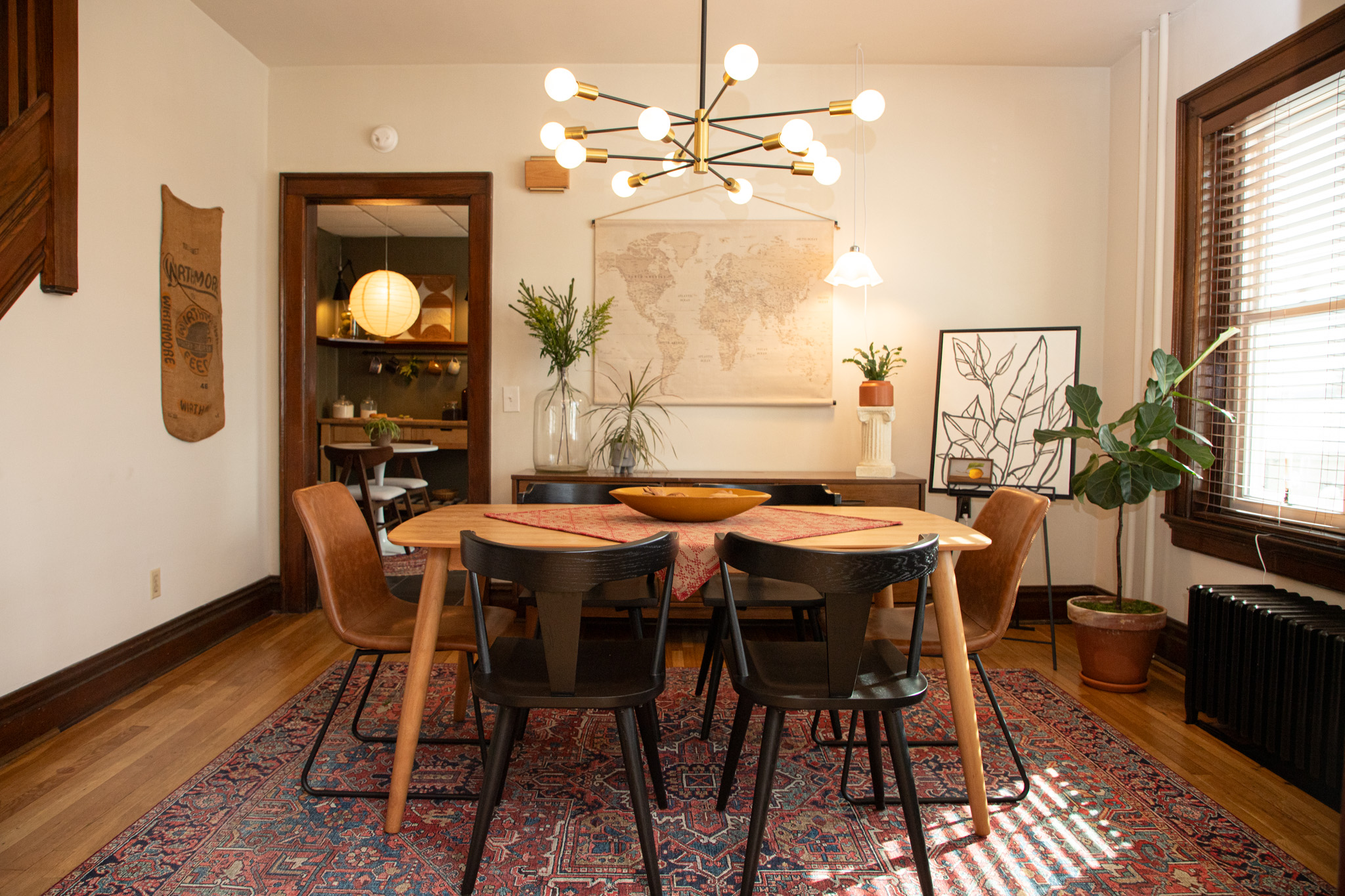Dining room with a wooden table, six chairs, a modern chandelier, and a large window with blinds, decorated with plants and artwork.