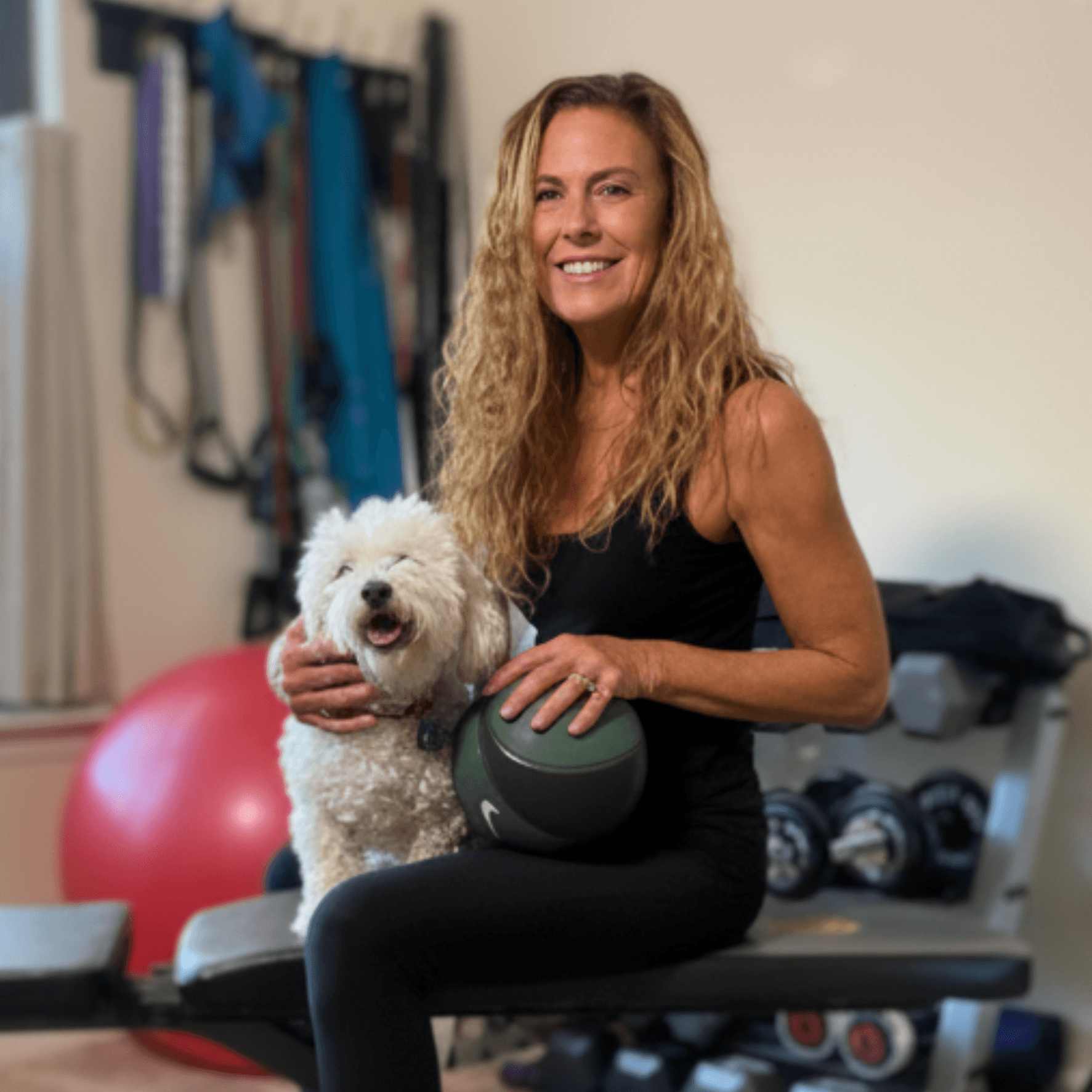 Woman with curly hair smiling, holding a white fluffy dog, in a fitness room with exercise equipment.