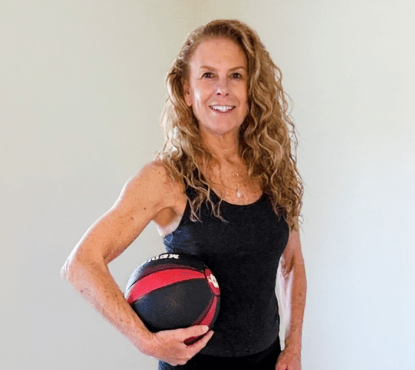 Woman with curly hair holding a medicine ball, smiling, wearing a black tank top, standing against a plain background.
