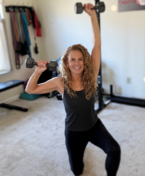Woman smiling, holding dumbbells, one arm raised, in a home gym with exercise equipment and hanging resistance bands