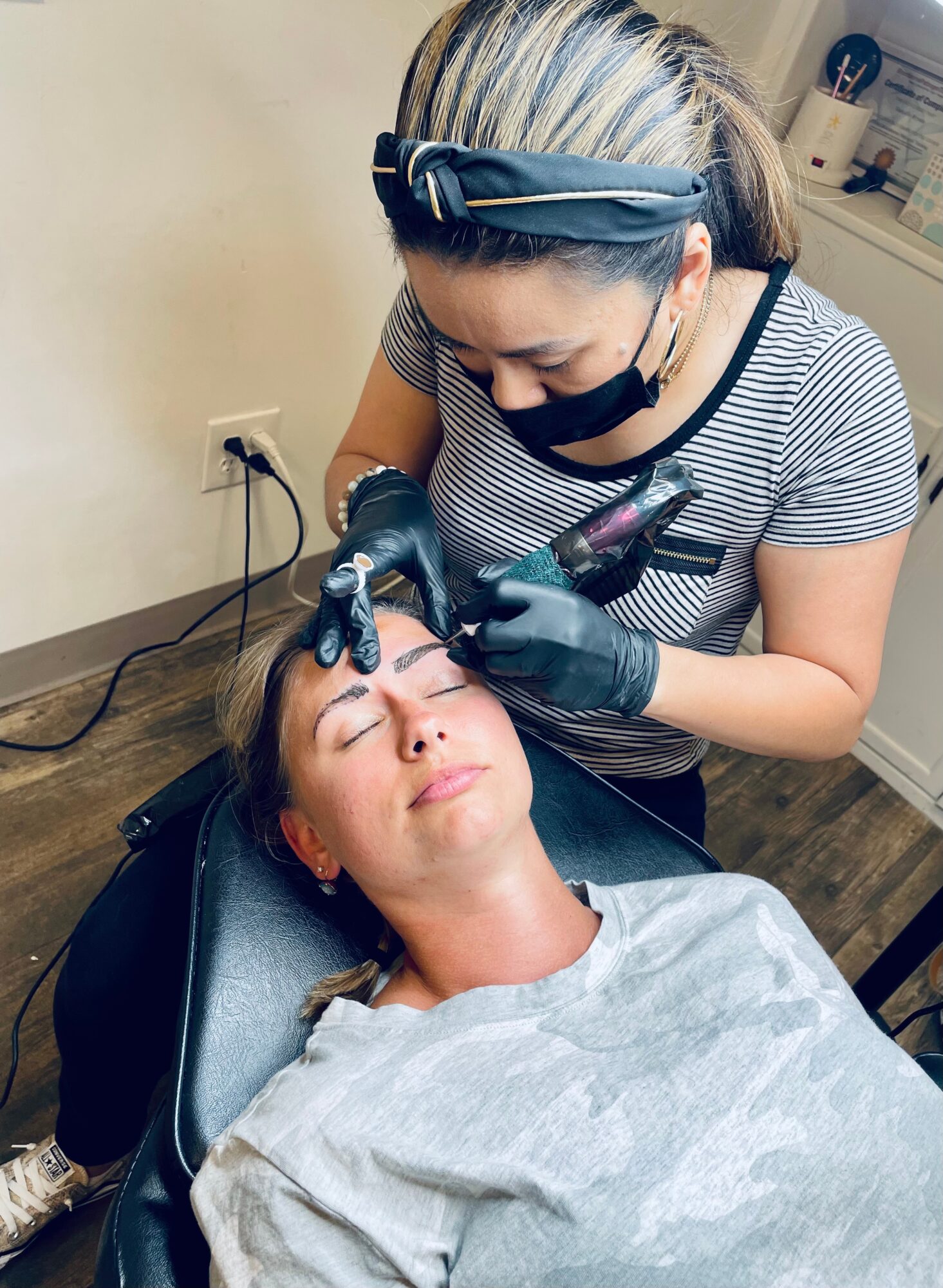A woman receives a cosmetic treatment from a professional in a clinic, lying back with eyes closed, wearing a gray cape.