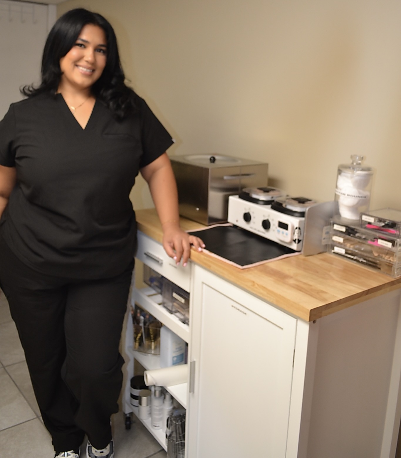 Woman in black scrubs standing next to a kitchen counter with appliances and storage, smiling, in a room with beige walls.