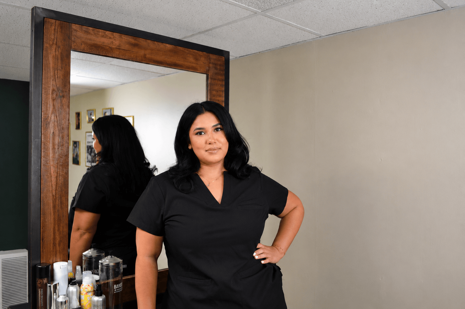 Woman with black hair and black shirt standing in front of mirror, hand on hip, in room with beige wall and ceiling tiles.