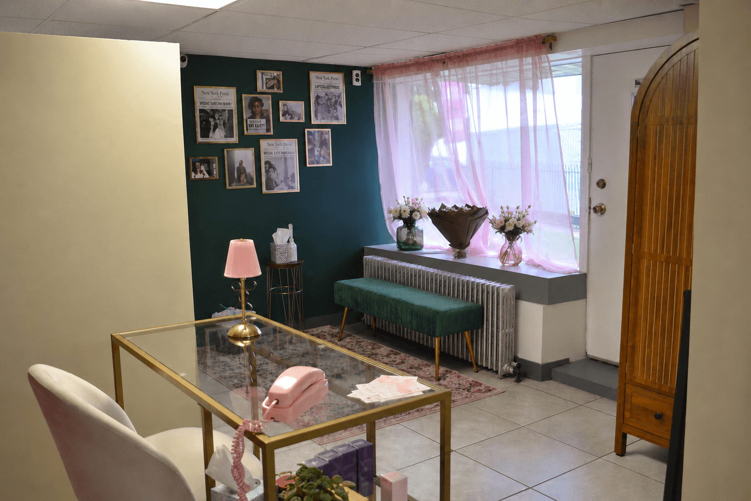 Living room with a green wall, framed photos, pink lamp, pink piggy bank, window with pink curtains, and a green bench.