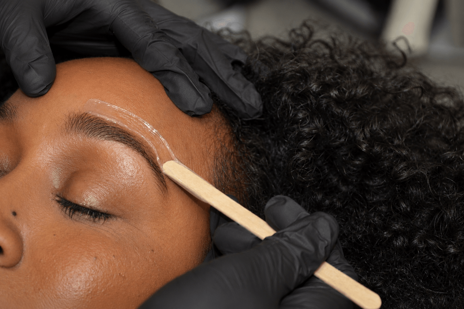 Person receiving eyebrow waxing or shaping, with a technician applying wax or product, close-up of face and hands, curly hair.