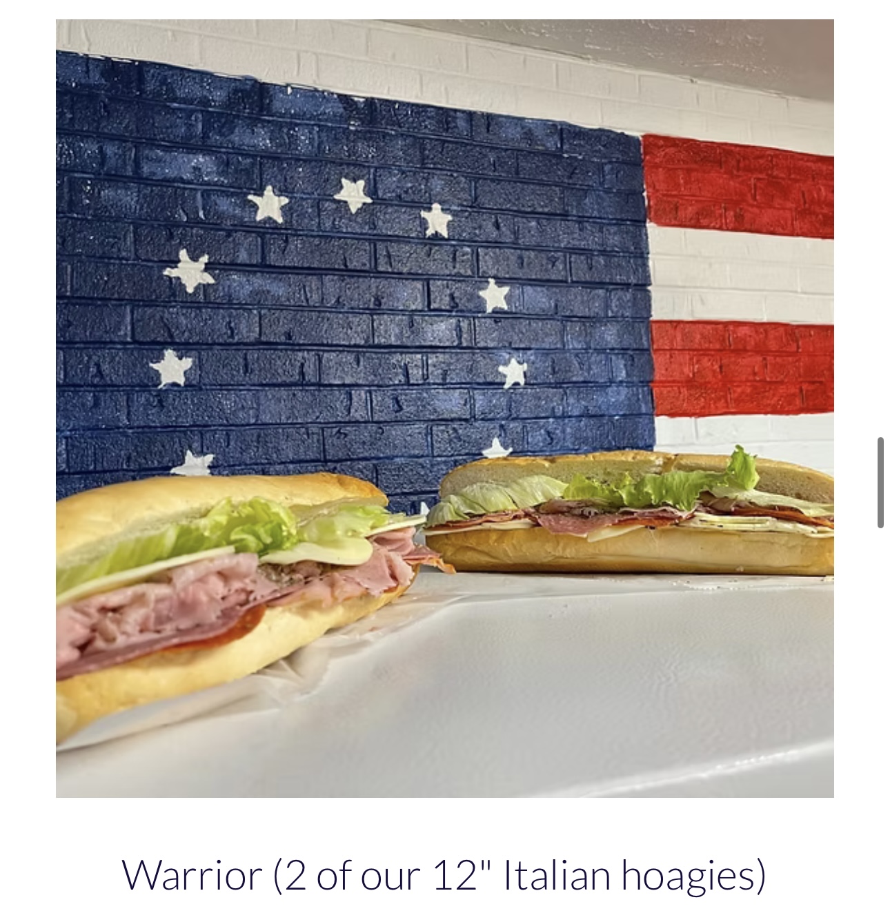 Two hoagies on a white surface with an American flag painted brick wall in the background.