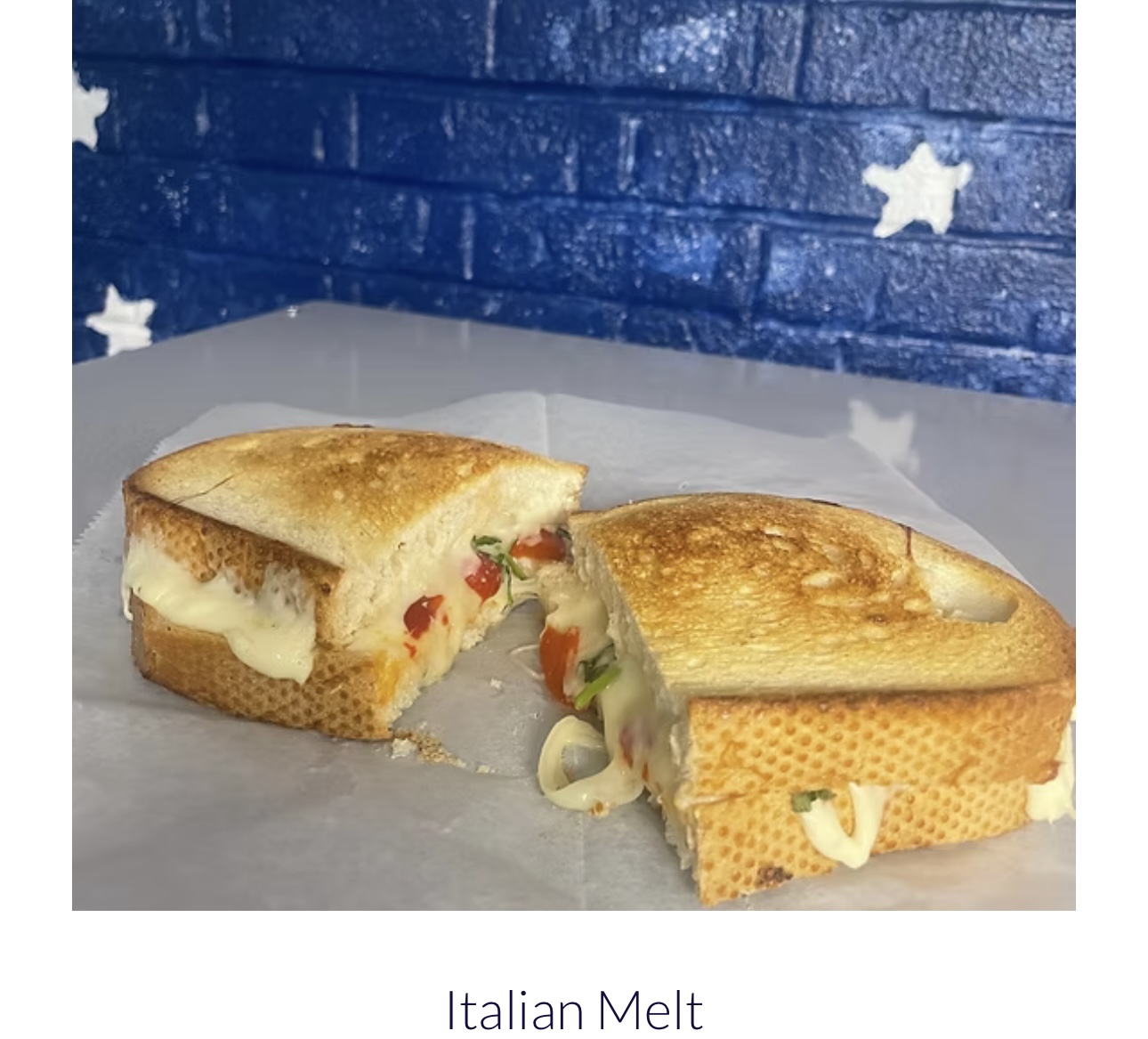 Sandwich with melted cheese, vegetables, and bread, cut in half, on white paper with a blue brick wall background.