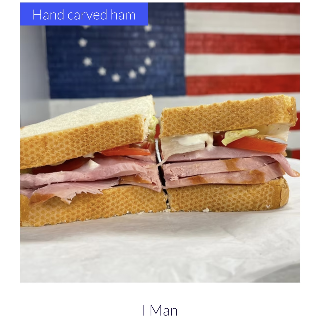 Two hand-carved ham sandwiches with ham, cheese, and tomato, placed on a surface with an American flag in the background.