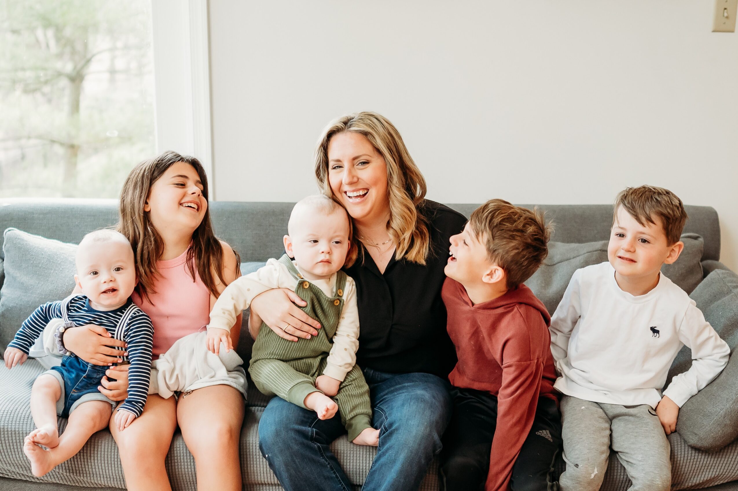Group of five people, including children and an adult woman, sitting on a couch indoors, smiling and interacting.