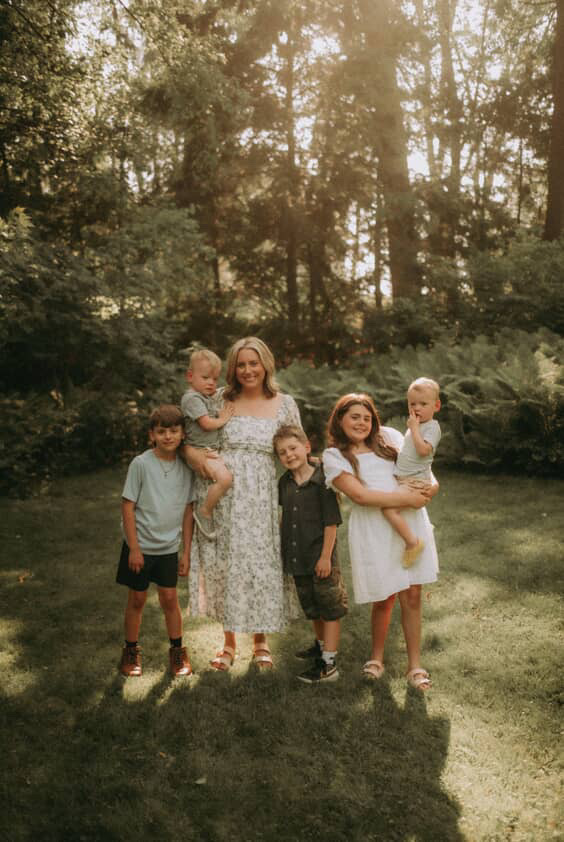 Group of five people, including children and an adult woman, standing outdoors on grass with trees in background.