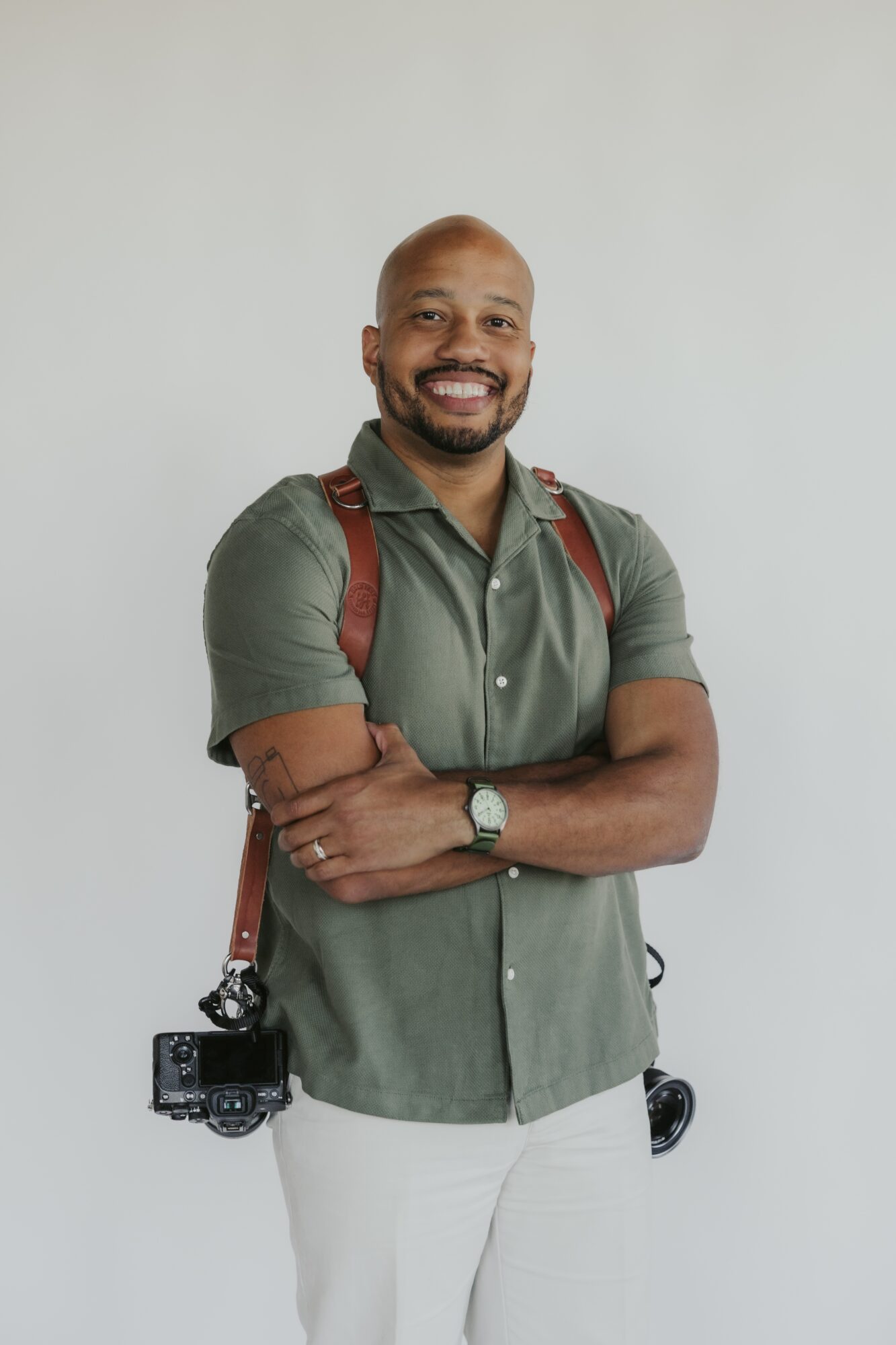 Smiling man with a beard, wearing a green shirt and white pants, standing with arms crossed against a plain background.