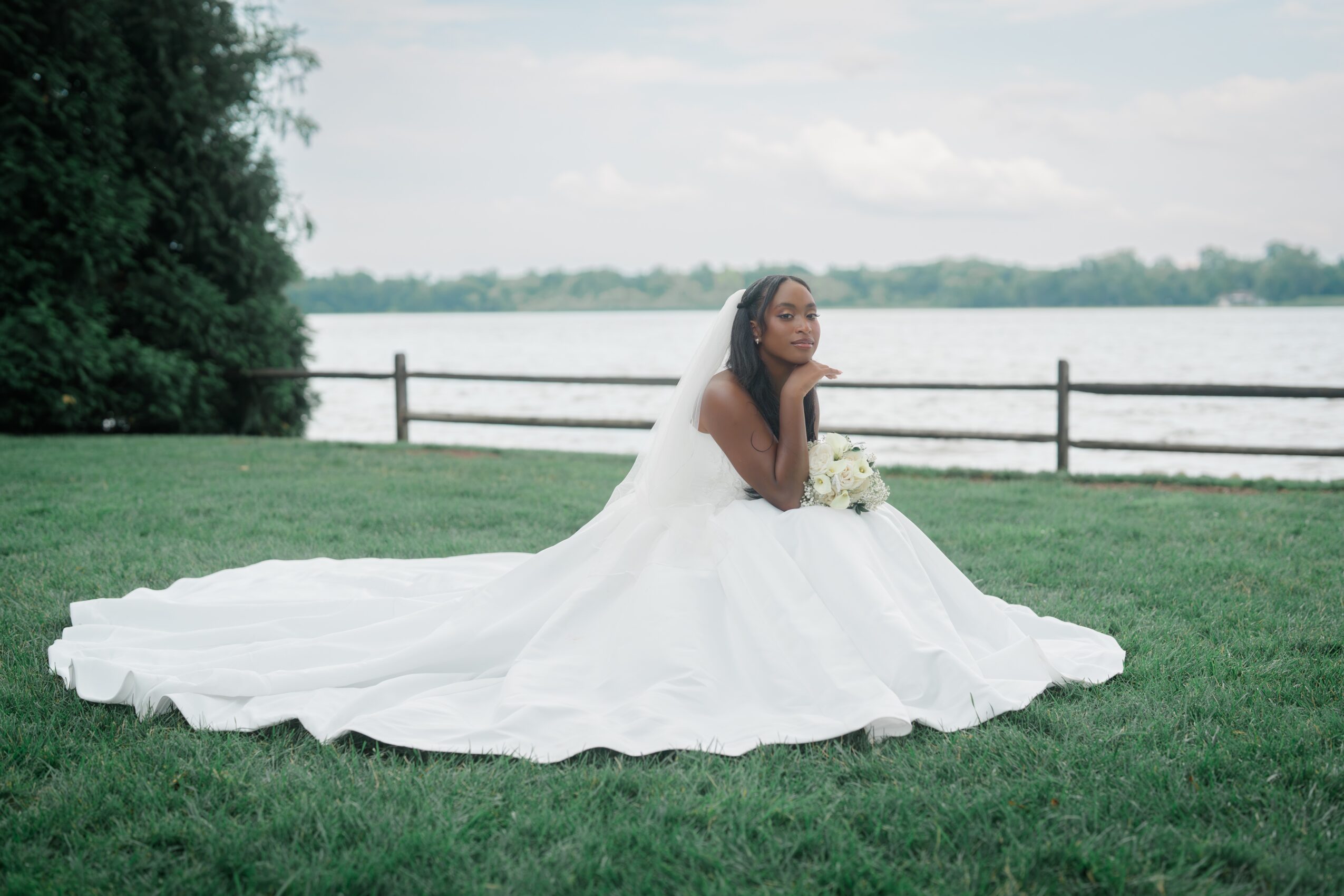 Woman in wedding dress sitting on grass near water with a fence and trees in background