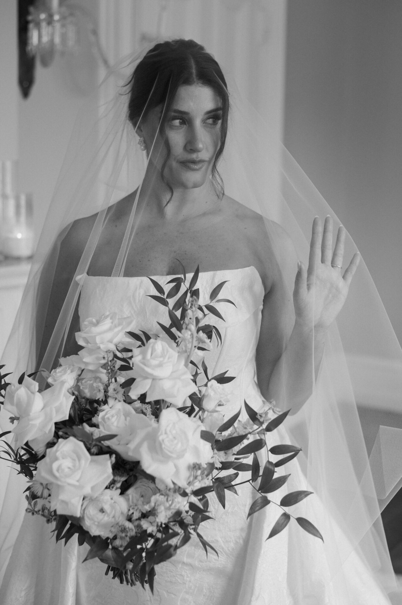 Bride holding a bouquet of flowers, wearing a strapless dress and veil, raising her hand, in a decorated room.