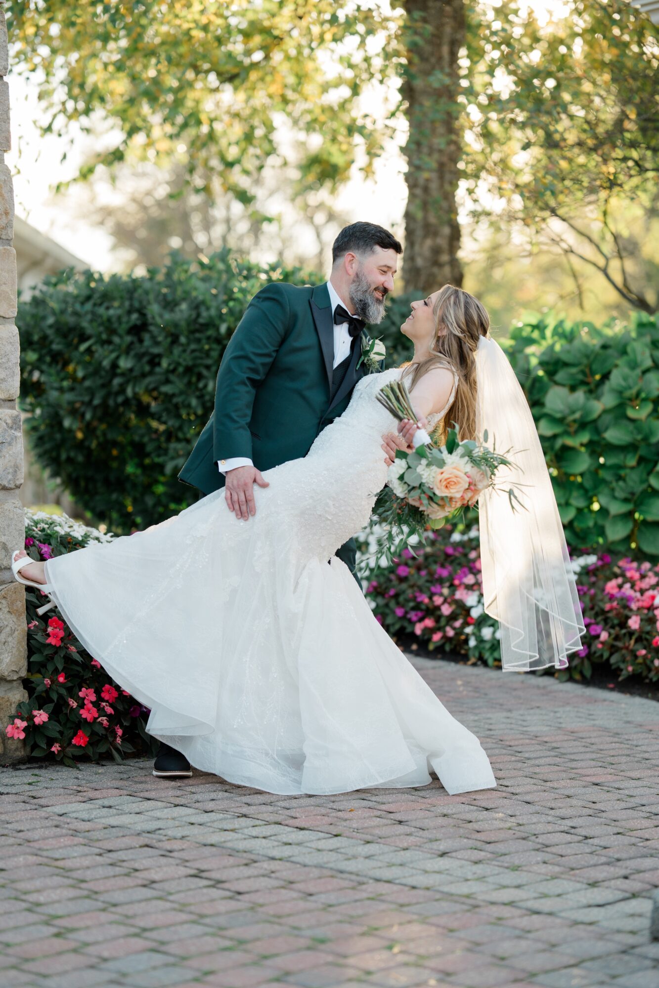 A bride and groom dance outdoors surrounded by greenery and flowers, with sunlight filtering through trees.