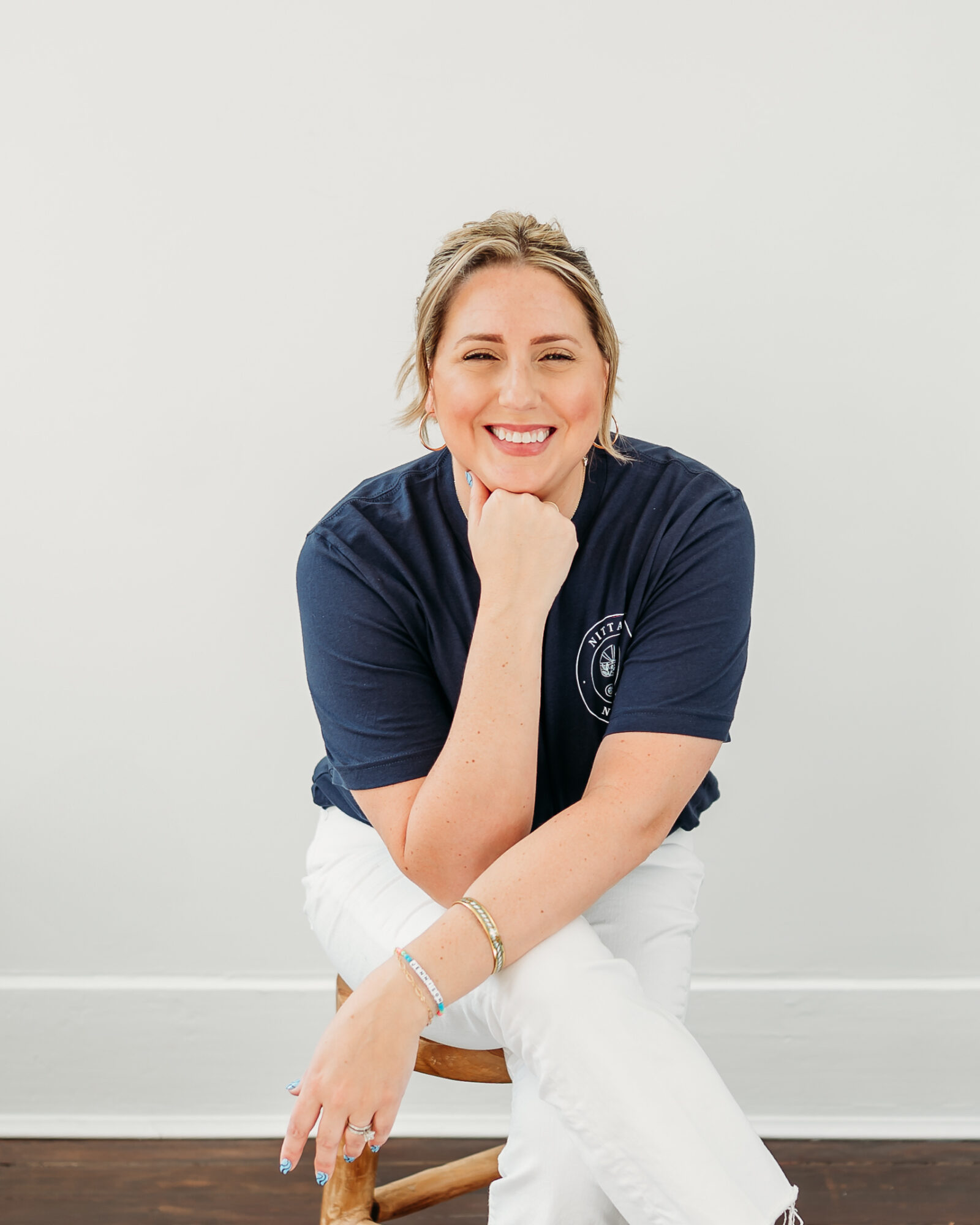 Smiling woman sitting on a stool, wearing a navy shirt and white pants, with her hand on her chin, against a plain background.