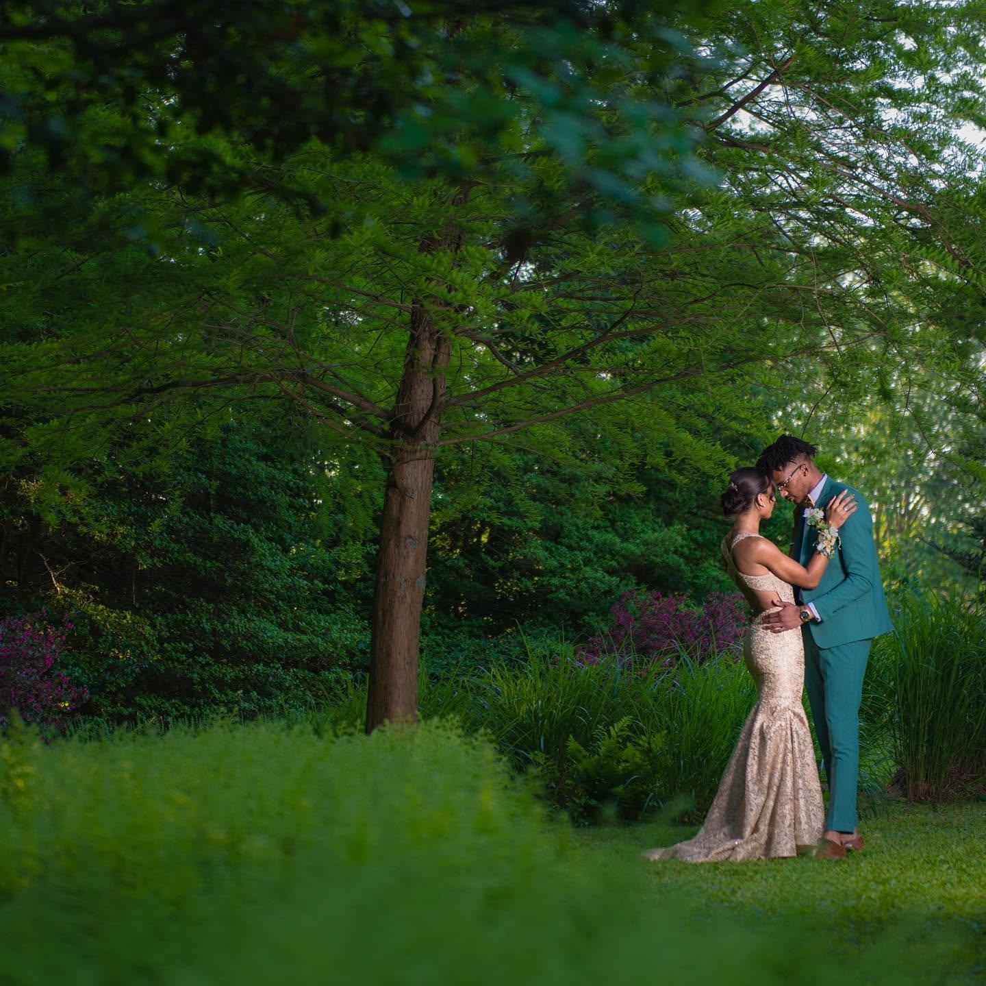 A couple in formal attire embracing in a lush green outdoor setting with trees and grass.