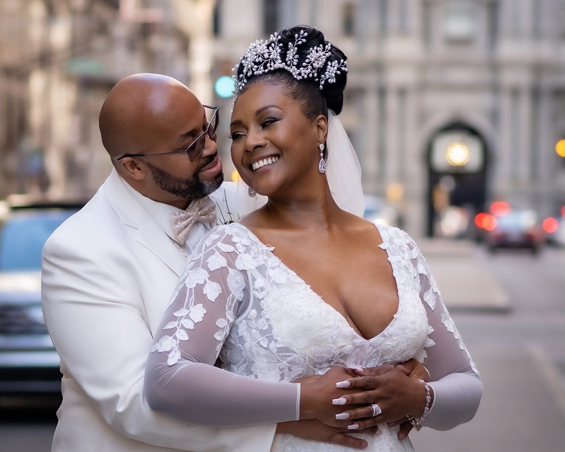 A smiling couple in wedding attire embracing outdoors in an urban setting, with buildings and cars in the background.