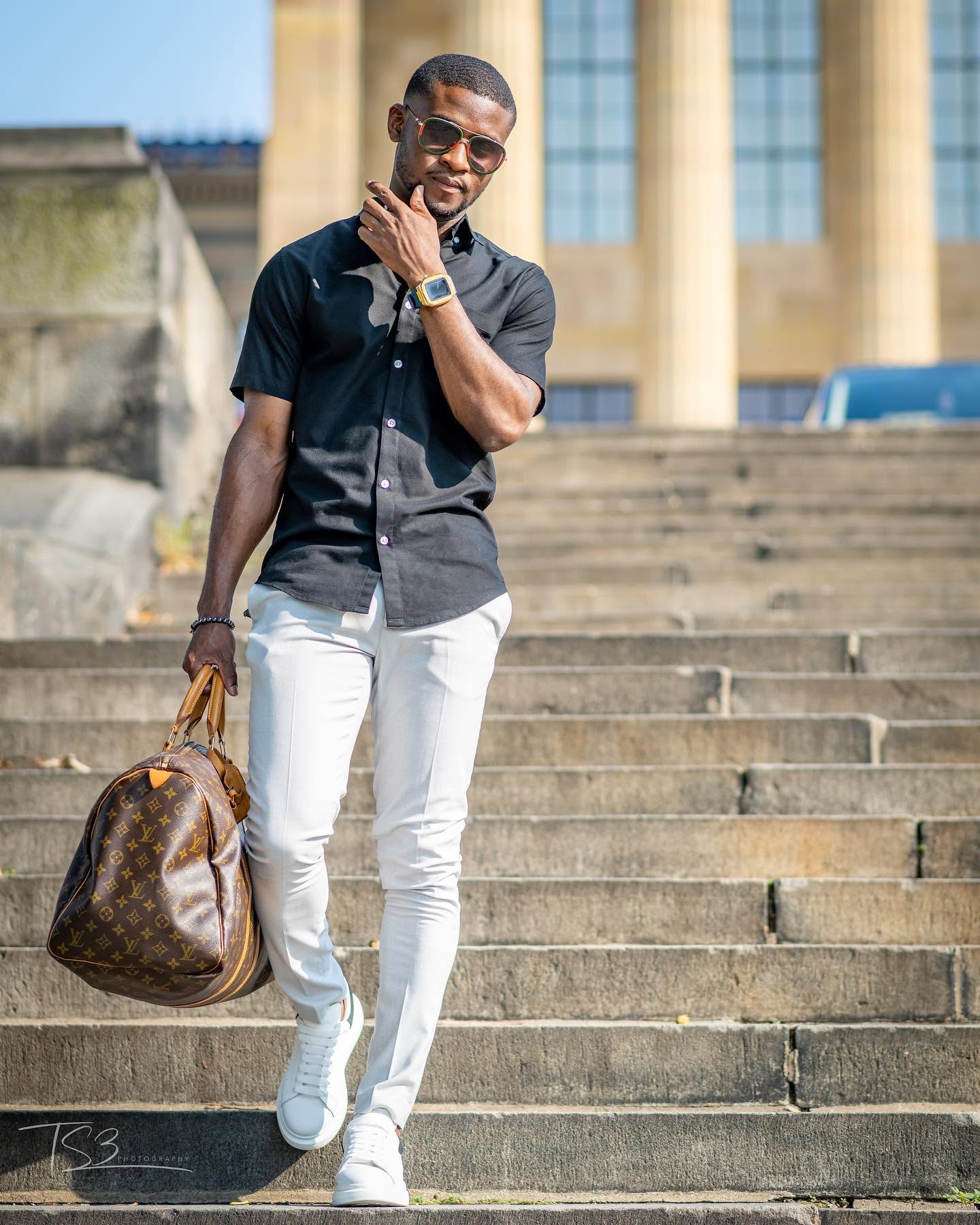 Man walking down stairs holding a bag, wearing sunglasses, black shirt, white pants, and a watch, outdoors in front of a building.