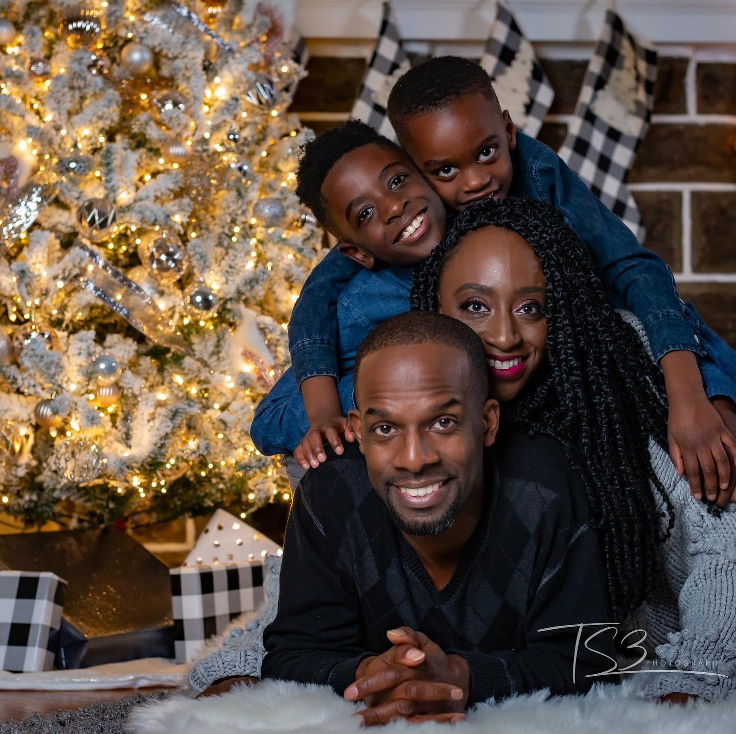 Family of four smiling together near a decorated Christmas tree, with presents and a fireplace in the background.