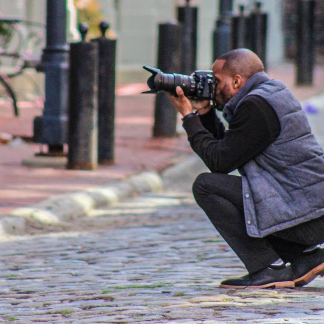 Person crouching on cobblestone street taking a photo with a camera, outdoors, with black posts and benches nearby.