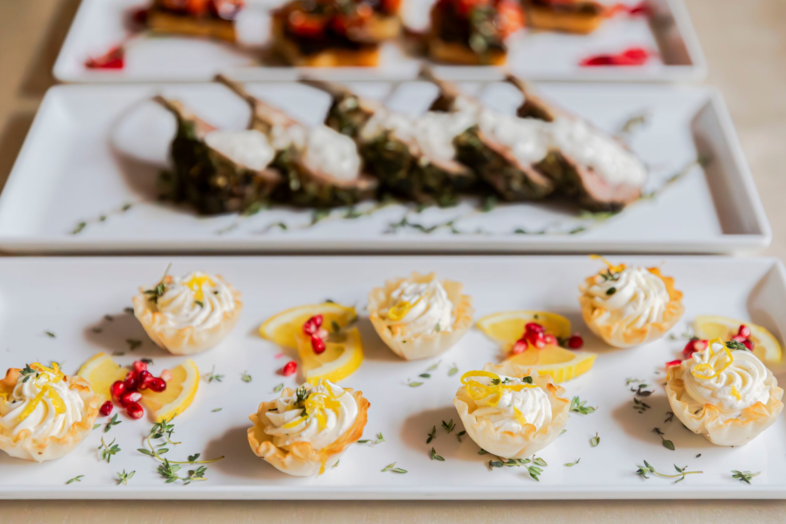 Assorted sushi and small desserts on white plates, garnished with herbs and red berries, with a blurred background.